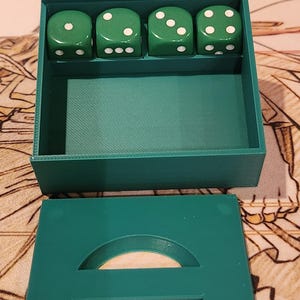 May include: A green dice box with four green dice. Each die has white dots representing numbers. The box has a lid with a circular cutout. The dice and box are on a patterned surface.
