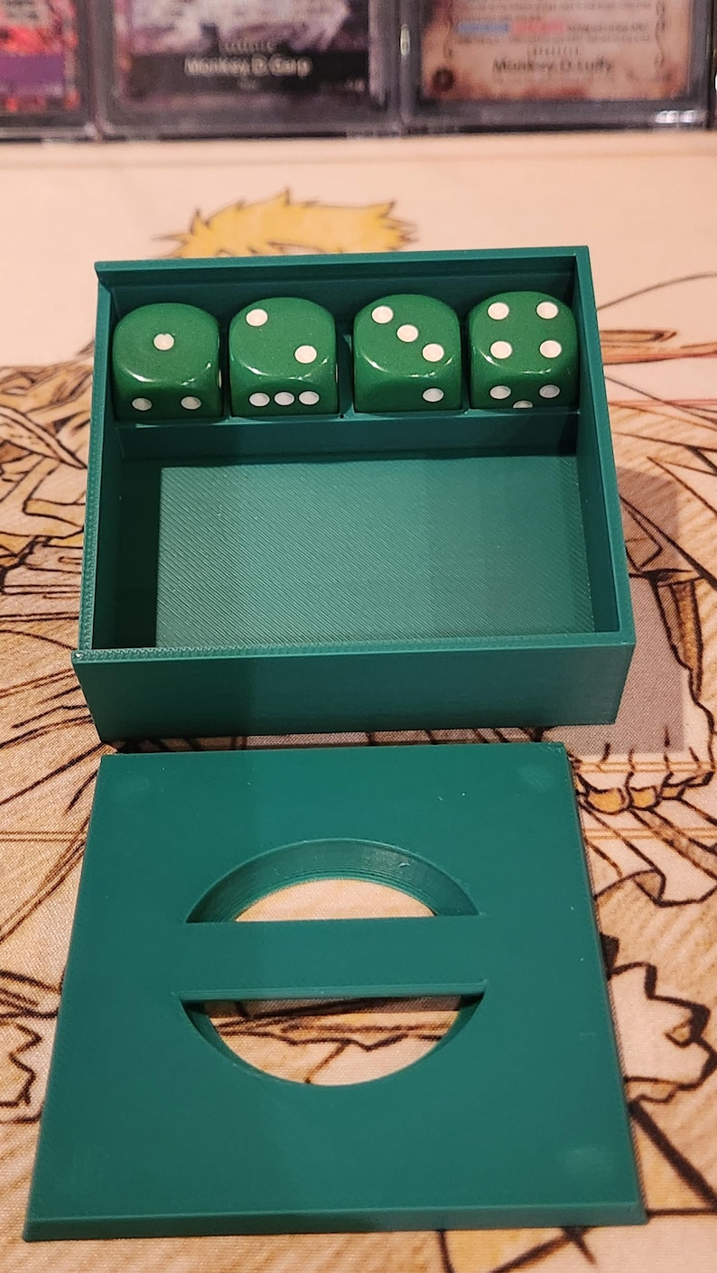May include: A green dice box with four green dice. Each die has white dots representing numbers. The box has a lid with a circular cutout. The dice and box are on a patterned surface.