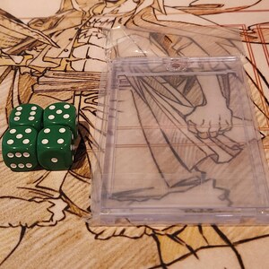 May include: Four green dice with white dots are arranged in a square, next to a clear plastic card holder. The background features a patterned surface with illustrations and text from trading cards.