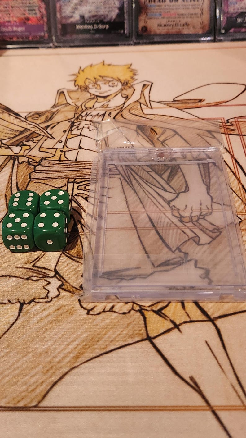 May include: Four green dice with white dots are arranged in a square, next to a clear plastic card holder. The background features a patterned surface with illustrations and text from trading cards.