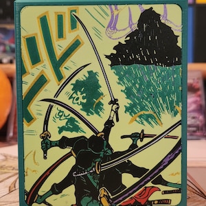 May include: A teal box featuring a dynamic illustration of sword-wielding figures in a battle scene. The artwork is in a Japanese woodblock print style, with a light green background and black and gold accents.