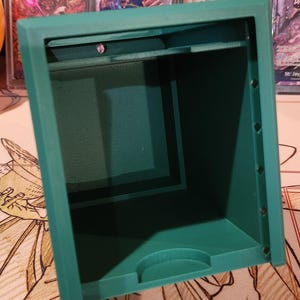 May include: A teal-colored, rectangular storage box with an open top. The box has a textured interior and is designed to hold small items. The box is sitting on a patterned surface, with trading cards visible in the background.