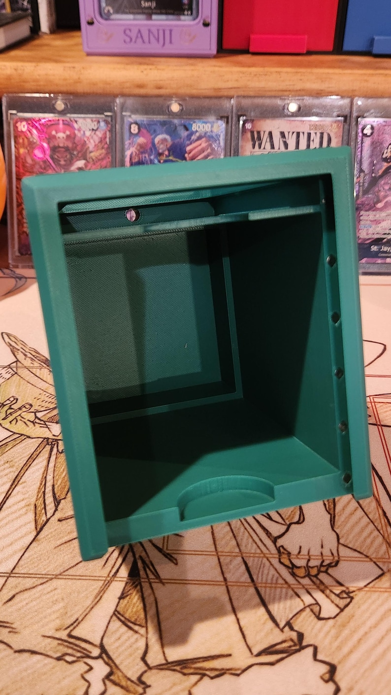 May include: A teal-colored, rectangular storage box with an open top. The box has a textured interior and is designed to hold small items. The box is sitting on a patterned surface, with trading cards visible in the background.