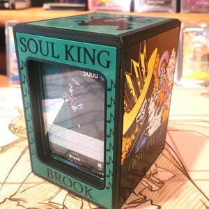 May include: A black and green card storage box with the text "SOUL KING" and "BROOK" in white. The box features a card with a skeleton character. The side of the box has a "ONE PIECE" graphic.