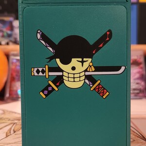 May include: A teal card storage box featuring a graphic of a skull with a black eye patch, crossed swords, and a black hat. The box is designed to hold playing cards or similar items. The design is a popular anime theme.