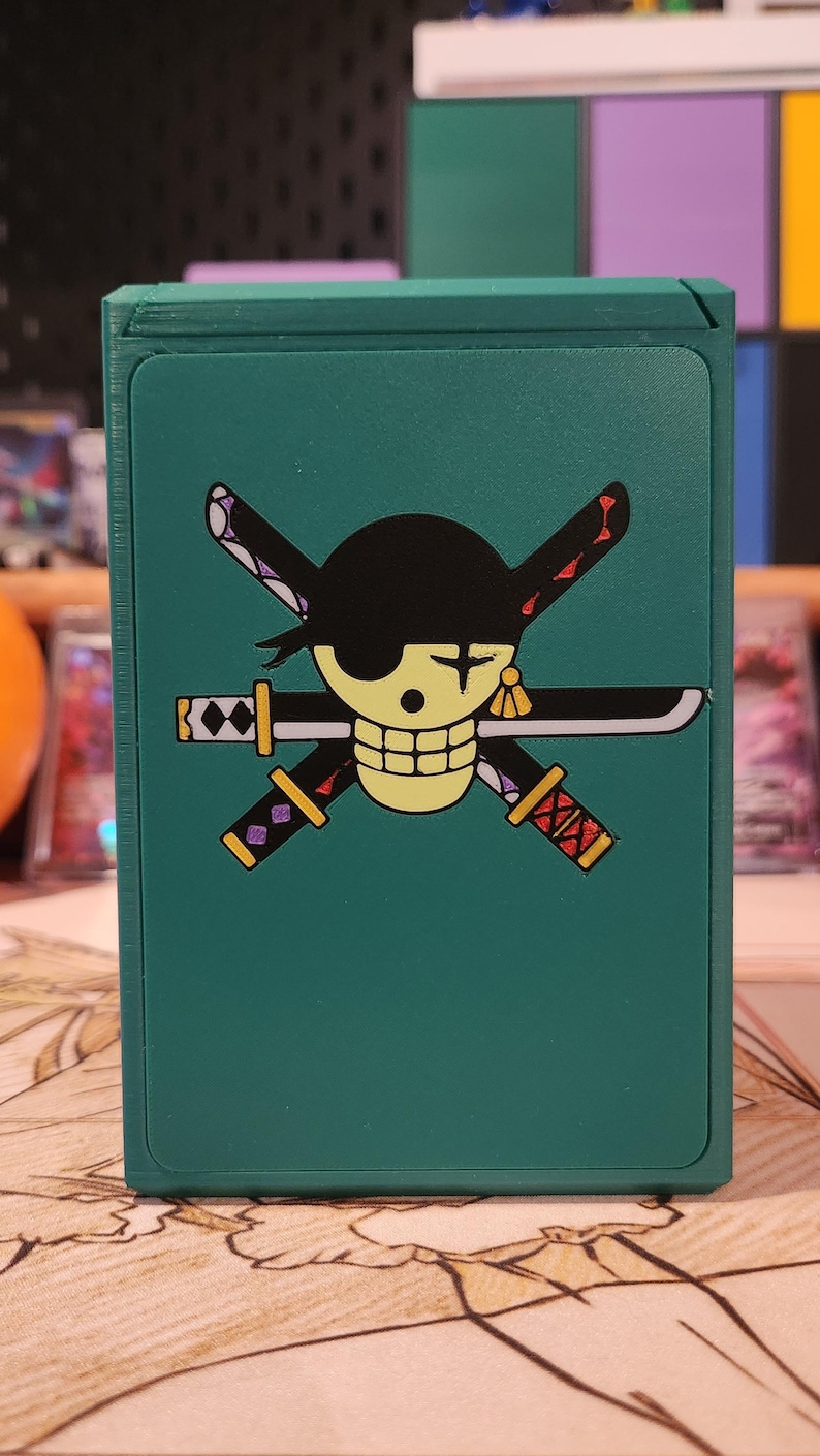 May include: A teal card storage box featuring a graphic of a skull with a black eye patch, crossed swords, and a black hat. The box is designed to hold playing cards or similar items. The design is a popular anime theme.