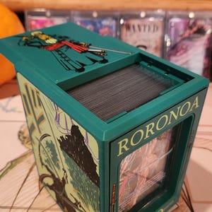 May include: A teal card storage box with the name "RORONOA" and "ZORO" printed on the sides. The box features illustrations of a character with swords and a top lid with a character illustration. The box is open, revealing a stack of cards.