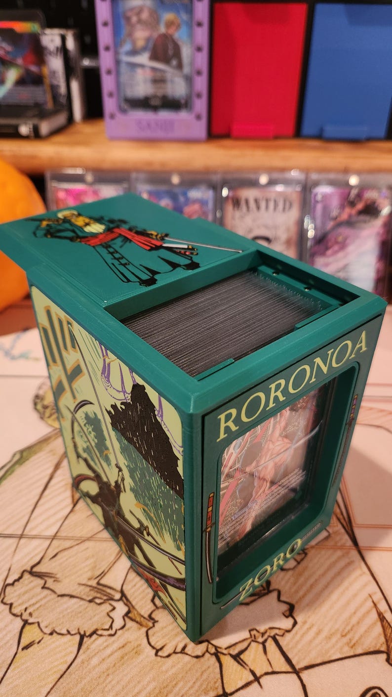 May include: A teal card storage box with the name "RORONOA" and "ZORO" printed on the sides. The box features illustrations of a character with swords and a top lid with a character illustration. The box is open, revealing a stack of cards.