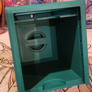 May include: A teal-colored, 3D-printed card storage box. The box has a rectangular shape with a recessed front panel featuring a stylized circular design. The interior is visible, and the box appears to be empty. The box is sitting on a patterned surface.