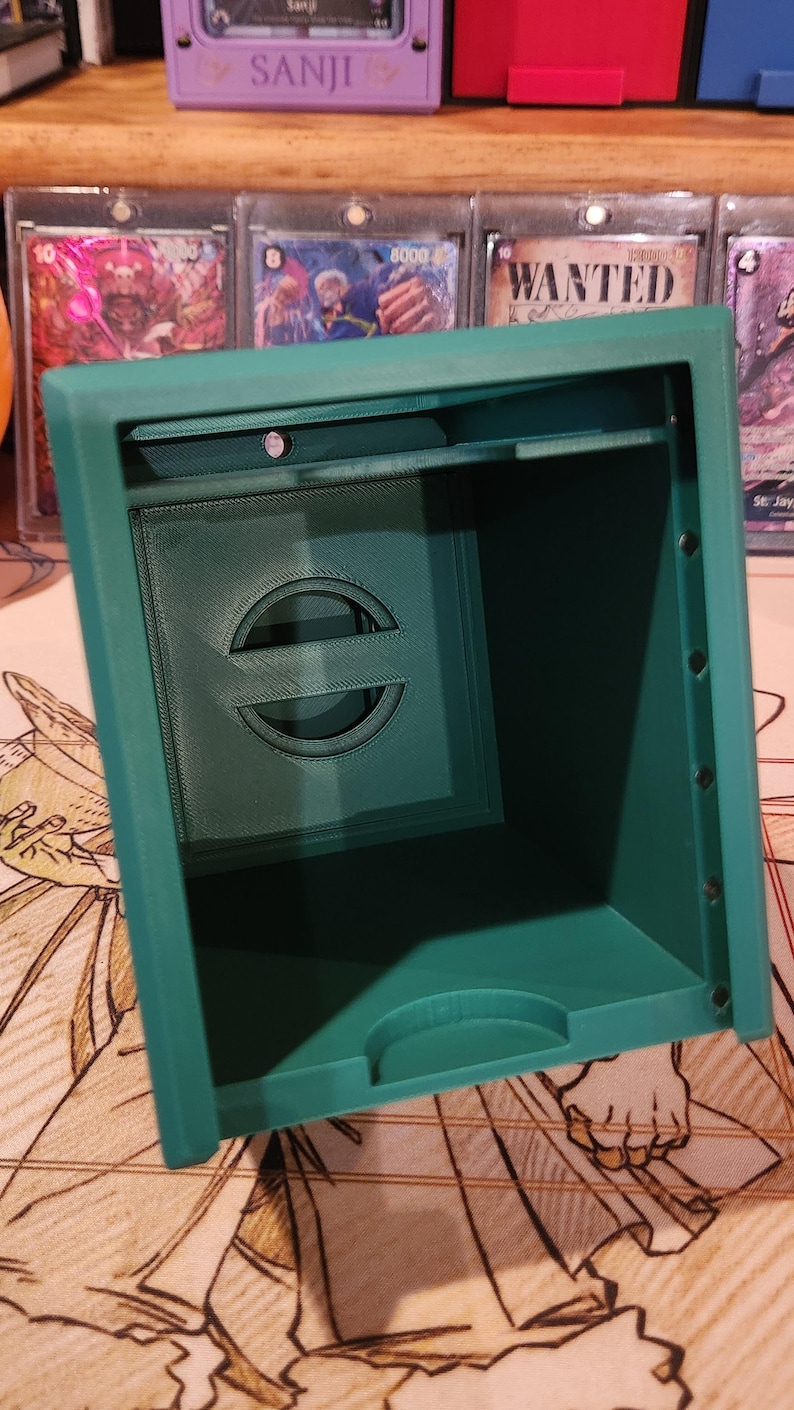 May include: A teal-colored, 3D-printed card storage box. The box has a rectangular shape with a recessed front panel featuring a stylized circular design. The interior is visible, and the box appears to be empty. The box is sitting on a patterned surface.
