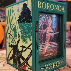 May include: A green card storage box featuring the name "RORONOA ZORO" and illustrations of a character with swords. The box has a clear window displaying a card with the same character. The box is on a patterned surface.