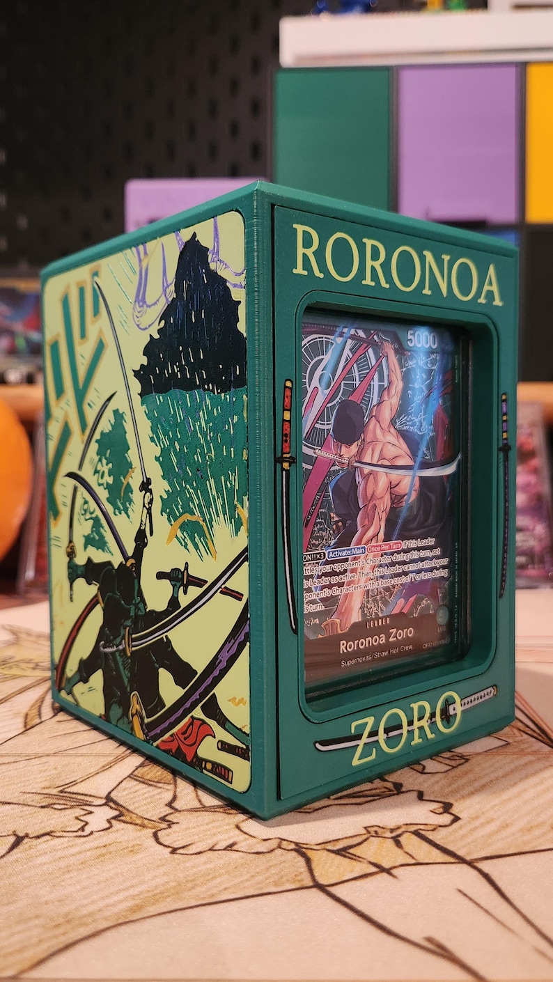 May include: A green card storage box featuring the name "RORONOA ZORO" and illustrations of a character with swords. The box has a clear window displaying a card with the same character. The box is on a patterned surface.