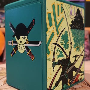 May include: A teal, square storage box featuring a skull and cross swords design on one side and a manga-style illustration of a swordsman on the other. The box has a hinged lid and is likely for storing collectibles.