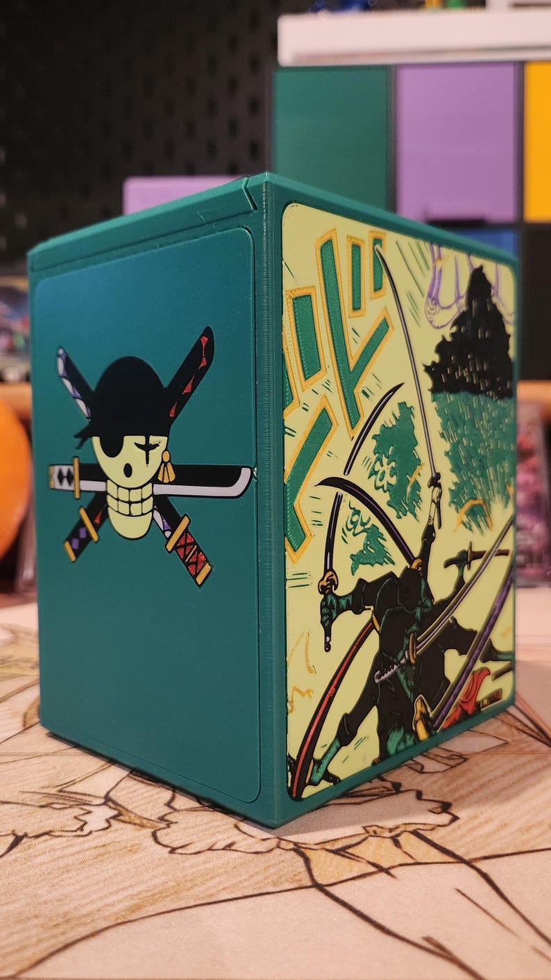 May include: A teal, square storage box featuring a skull and cross swords design on one side and a manga-style illustration of a swordsman on the other. The box has a hinged lid and is likely for storing collectibles.