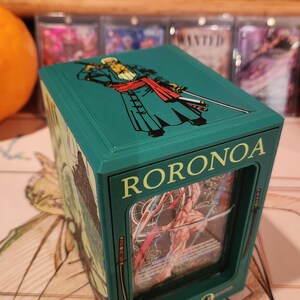 May include: A teal card storage box featuring the character Roronoa Zoro from the One Piece anime. The box has a clear window displaying a card, with the name "RORONOA" and "ZORO" printed on the front.