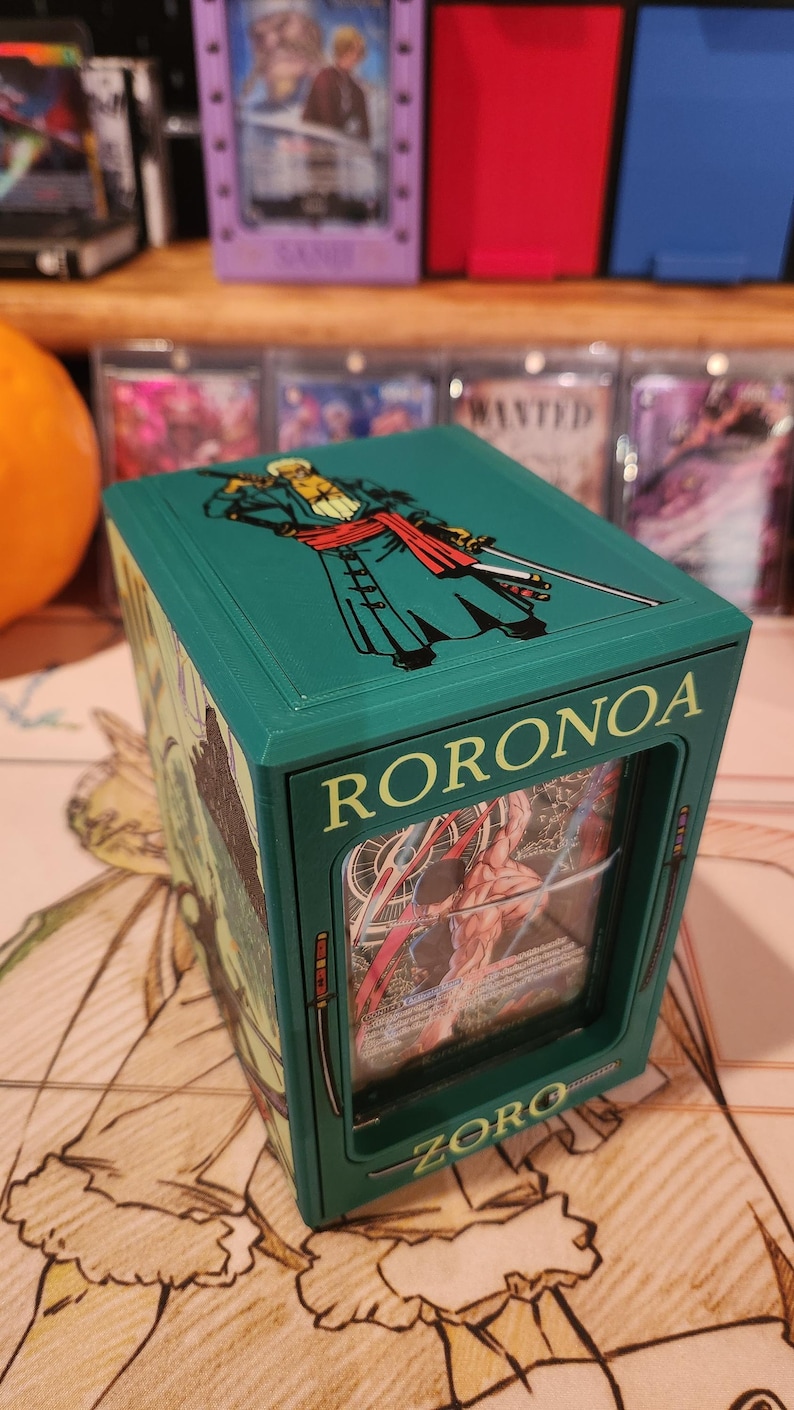 May include: A teal card storage box featuring the character Roronoa Zoro from the One Piece anime. The box has a clear window displaying a card, with the name "RORONOA" and "ZORO" printed on the front.