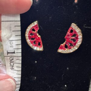 May include: A pair of watermelon slice earrings featuring red enamel centers with black seed cutouts and clear rhinestone borders. A white measuring tape is positioned next to the earrings, showing they are approximately 0.75 inches in height.