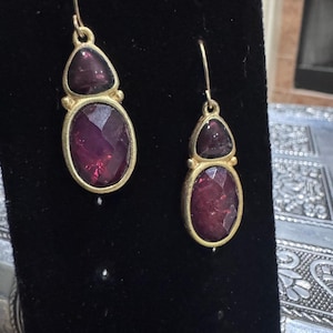 May include: A pair of gold-toned earrings with a deep red, faceted gemstone design. Each earring features a smaller, triangular stone above a larger, oval-shaped stone. The earrings are displayed against a black background.