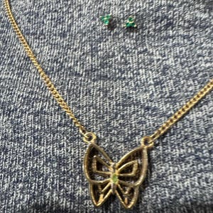 May include: A gold-toned butterfly pendant necklace with a delicate chain. The butterfly has an openwork design and a small green gem in the center. Matching green gem stud earrings are also visible.