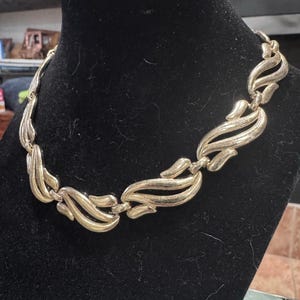May include: A gold-toned necklace with a repeating, stylized leaf design. The necklace is displayed on a black velvet bust, showcasing its intricate details and vintage style. The links are connected, forming a flexible chain.