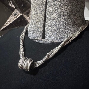 May include: A silver-toned necklace with a braided design. The necklace features a central, cylindrical knot detail. The jewelry is displayed on a black background, showcasing the intricate metalwork and texture of the piece.