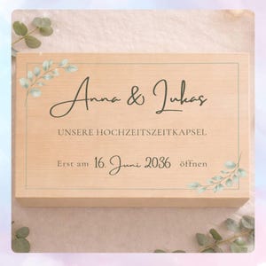 Könnte beinhalten: Eine hellfarbene Holzkiste mit den Namen "Anna & Lukas" in eleganter Schrift. Der Text "UNSERE HOCHZEITSZEITKAPSEL" und "Erst am 16. Juni 2036 öffnen" sind ebenfalls sichtbar. Die Kiste ist mit aquarellartigen Grünpflanzen verziert.
