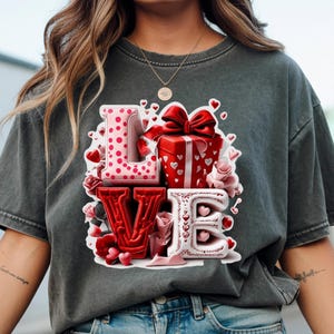 Peut inclure: T-shirt gris foncé avec le mot "LOVE" en design 3D. Le "L" est rose à pois blancs, le "V" est rouge et le "E" est blanc avec des cœurs rouges. Une boîte cadeau rouge et des roses sont également présentes.