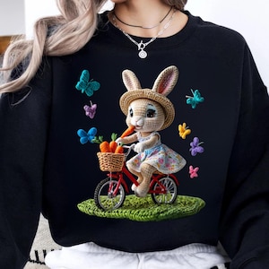 Peut inclure: Sweat-shirt noir avec un lapin sur un vélo rouge, portant des carottes dans un panier. Le lapin porte un chapeau de paille et une robe à fleurs. Des papillons colorés entourent le lapin. Illustration fantaisiste.