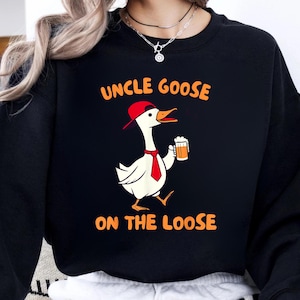 Peut inclure: Sweat-shirt noir avec l'inscription "UNCLE GOOSE ON THE LOOSE". Le graphisme représente une oie de dessin animé portant une casquette et une cravate rouges, tenant une chope de bière. L'oie est blanche avec un bec et des pieds orange.