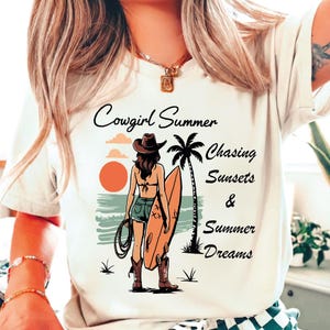 Peut inclure: Un t-shirt couleur crème avec un motif représentant une personne portant un chapeau de cow-boy, un haut de bikini, un short en jean et des bottes de cow-boy, tenant une planche de surf et un lasso. Le texte indique Cowgirl Summer Chasing Sunsets & Summer Dreams.