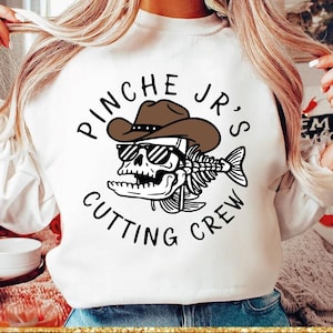 Peut inclure: Sweat-shirt blanc avec un motif représentant un crâne portant un chapeau de cowboy et des lunettes de soleil, avec un squelette de poisson et le texte "PINCHE JR'S CUTTING CREW" dans un design circulaire. Le motif est en noir et marron.