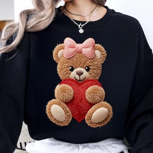 Peut inclure: Sweat-shirt noir avec un ours en peluche brodé tenant un cœur rouge. L'ours en peluche a un nœud rose et est fait de tissu marron texturé. Un design mignon et confortable pour un look décontracté.