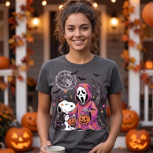 Peut inclure: Un t-shirt gris foncé avec une impression graphique représentant un beagle blanc portant un chapeau de sorcière violet, à côté d'une silhouette en robe violette à capuche et masque de fantôme blanc, tenant des seaux citrouilles orange.