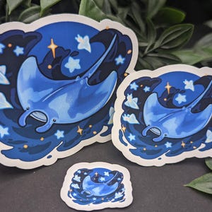 May include: Three stickers featuring a blue manta ray design. The manta rays are depicted with a dark blue background, surrounded by stars and clouds. The stickers vary in size, with the largest measuring approximately 4 inches wide.