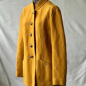 Könnte beinhalten: Senfgelbe Wolljacke mit Stehkragen und drei dekorativen Knöpfen. Die Jacke hat eine taillierte Passform, eine kleine Tasche und ein silberfarbenes Emblem. Das Kleidungsstück wird auf einer Schneiderpuppe präsentiert.