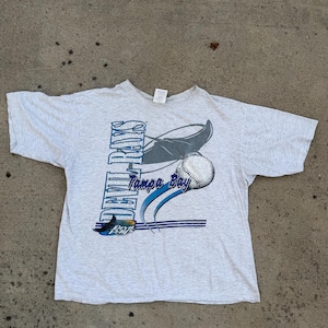 Puede incluir: Camiseta gris claro con un diseño gráfico. El diseño incluye las palabras "Tampa Bay" en azul y blanco, una pelota de béisbol y un bate de béisbol estilizado. La camiseta está hecha de un material suave.