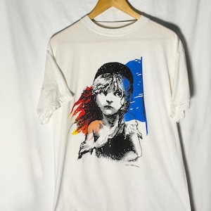 May include: White t-shirt with a graphic of a young person wearing a black hat, with a blue, red, and yellow design. The graphic is a stylized illustration. The shirt is hanging on a wooden hanger.