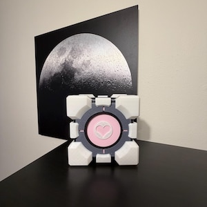 May include: A white, grey, and pink Companion Cube, a fictional item from the video game Portal, sits on a black surface. The cube features a heart symbol in the centre. A black and white moon print hangs in the background.