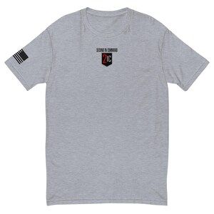 Puede incluir: Una camiseta gris jaspeado con una pequeña bandera estadounidense en la manga izquierda. La camiseta tiene el texto "SECOND IN COMMAND" sobre un escudo rojo y negro con "2IC" impreso en el pecho.