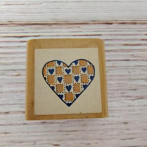 May include: A square wooden rubber stamp featuring a heart-shaped design on its top surface. The heart is filled with a repeating pattern of small blue hearts and light brown squares, outlined in a dark blue border on a cream-colored background.
