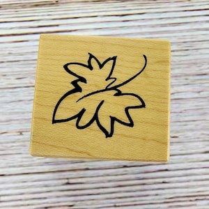 May include: A square, wooden stamp with a black outline of a leaf design. The stamp is made of light-colored wood and sits on a light-colored, wooden surface. The leaf design is a simple, stylized illustration.