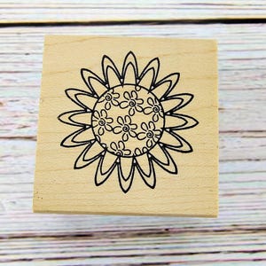 May include: A wooden stamp with a black outline of a sunflower design. The sunflower has a detailed center with small flower shapes and large petals. The stamp is square and sits on a light wood surface.