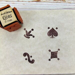 May include: A brown rubber stamp with the text "Jedi Kins Obits ©1996" next to four stamped playing card suit symbols in dark brown ink on a cream-colored paper. The symbols include a club, spade, a swirl, and a diamond.