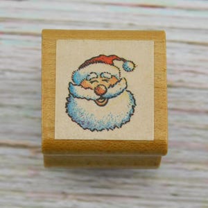 May include: A wooden stamp featuring a smiling Santa Claus face with a red hat, blue beard, and rosy cheeks. The stamp is rectangular and has a light brown wooden base. The Santa image is printed on a square piece of paper.