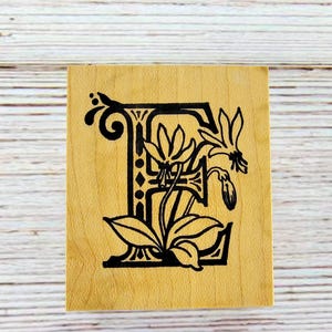 May include: Wooden stamp featuring the letter "E" in a decorative, black floral design. The stamp is square and made of light-colored wood. The floral design includes leaves and flowers.