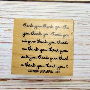May include: A wooden stamp with the words "thank you" repeated in a handwritten style. The stamp is square and has the text "© 2004 STAMPIN' UP!" at the bottom. The stamp is on a wooden surface.