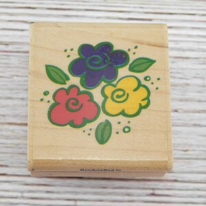 May include: Wooden stamp featuring a colorful floral design. The stamp has three flowers in red, yellow, and purple, with green leaves. The stamp is on a light wood block.