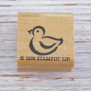 May include: A wooden stamp featuring a simple black outline of a duck. The stamp has the text "© 1998 STAMPIN' UP!" printed in black. The stamp is square and made of light-colored wood.