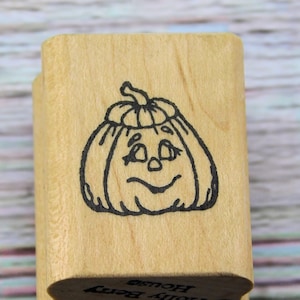 May include: A rectangular wooden rubber stamp featuring a black line art illustration of a pumpkin with a smiling face. The light-colored wood block has the text Holly Berry House stamped in black ink along the bottom edge of the side surface.