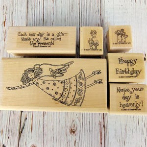 May include: A collection of five wooden rubber stamps featuring black ink designs. The largest stamp depicts a flying angel. Other stamps include text reading Each new day is a gift that's why it's called the present, Happy Birthday, and Hope your day is heavenly, plus a flower and a gift box.